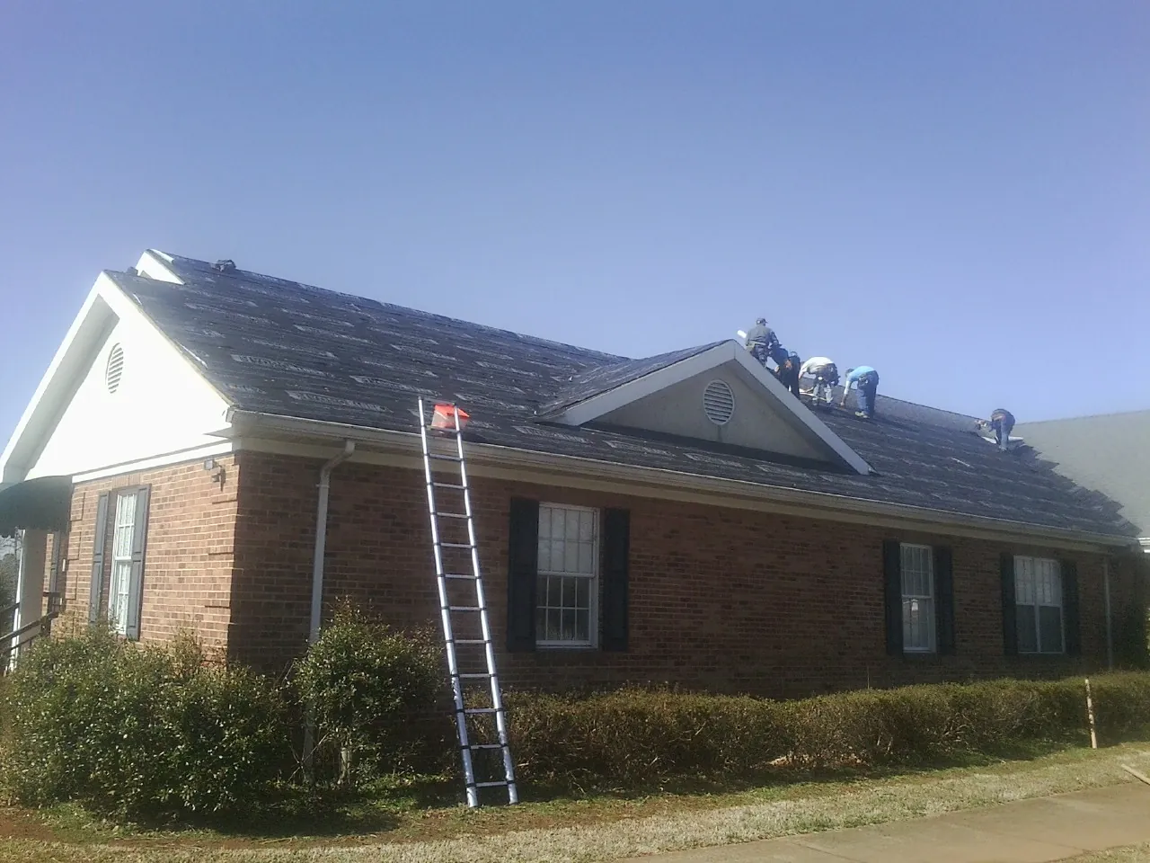 Crew tearing shingles off a brick ranch roof with a ladder set — Emergency Roof Repair work by our crew in Hasty
