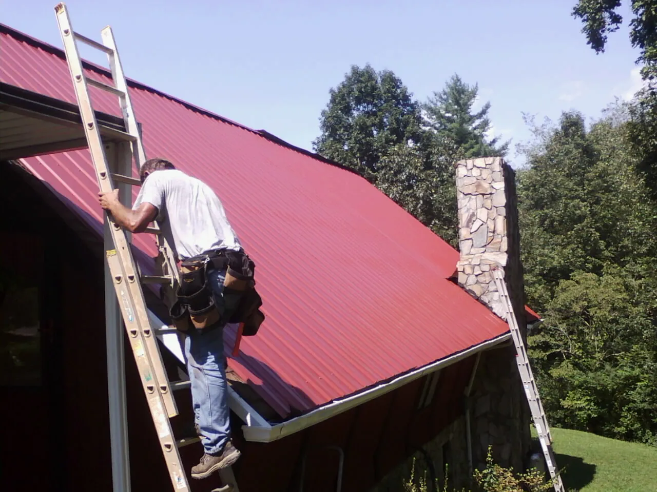 Roofer on a ladder beside a red metal roof and stone chimney — Soffit & Fascia Repair by Roofing Brothers in Hasty
